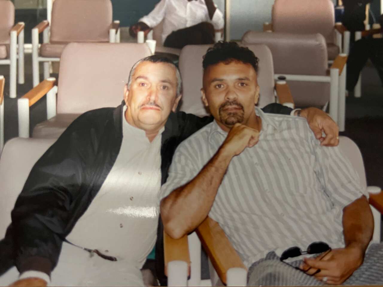 Two men sitting, one with his arm around the other