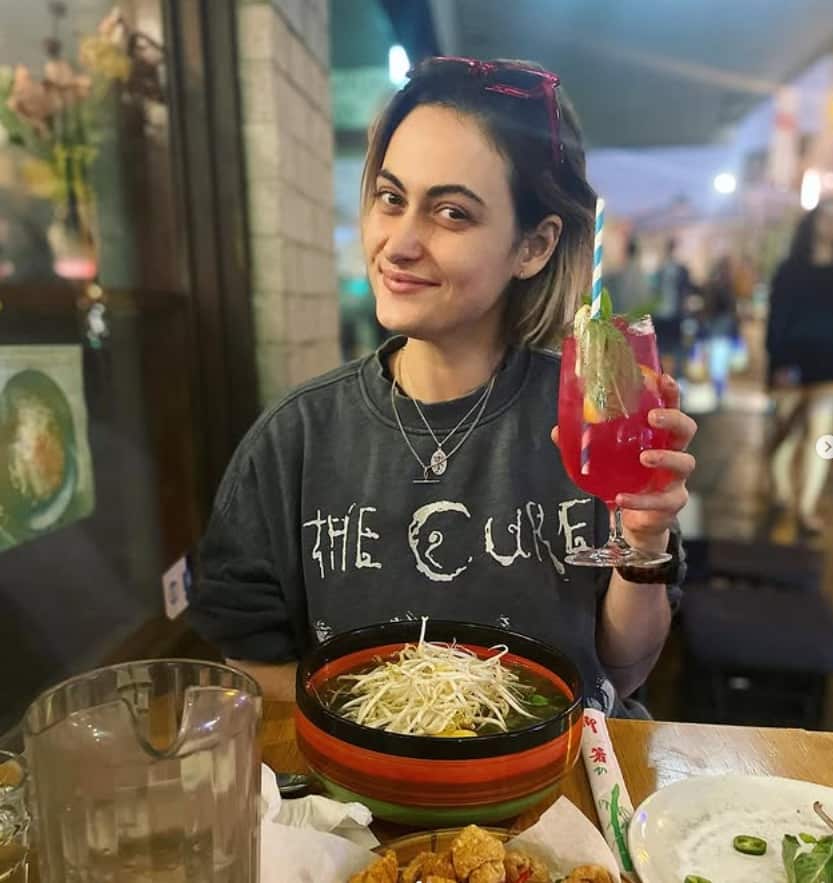 A woman in a Cure T-shirt is eating pho and drinking a cocktail. She is raising her glass and smiling at the camera.
