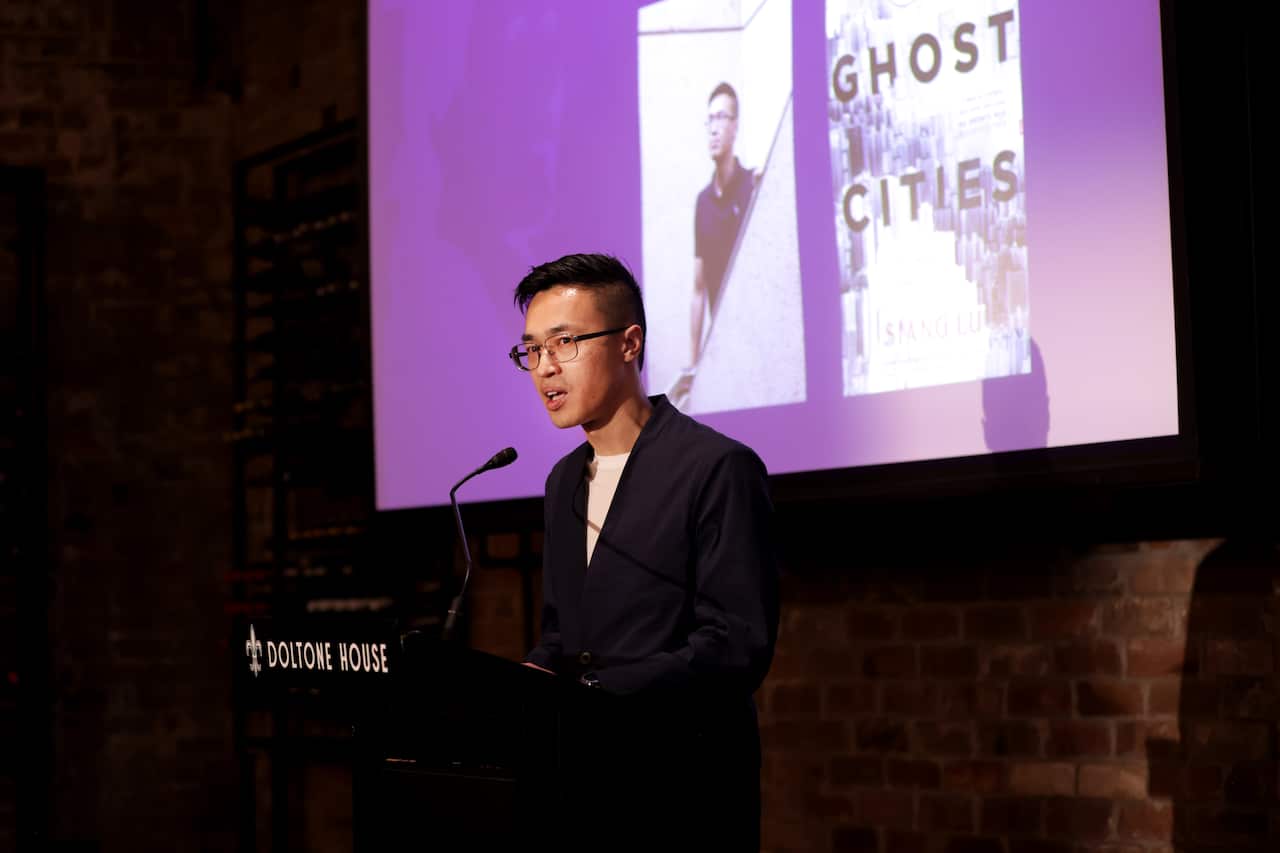 A man speaking at a podium into a microphone. On a screen behind him, a book called "Ghost Cities" is displayed.