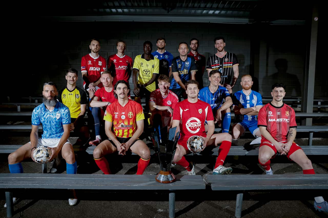 Footballers in different coloured jerseys sitting on a bench behind a trophy