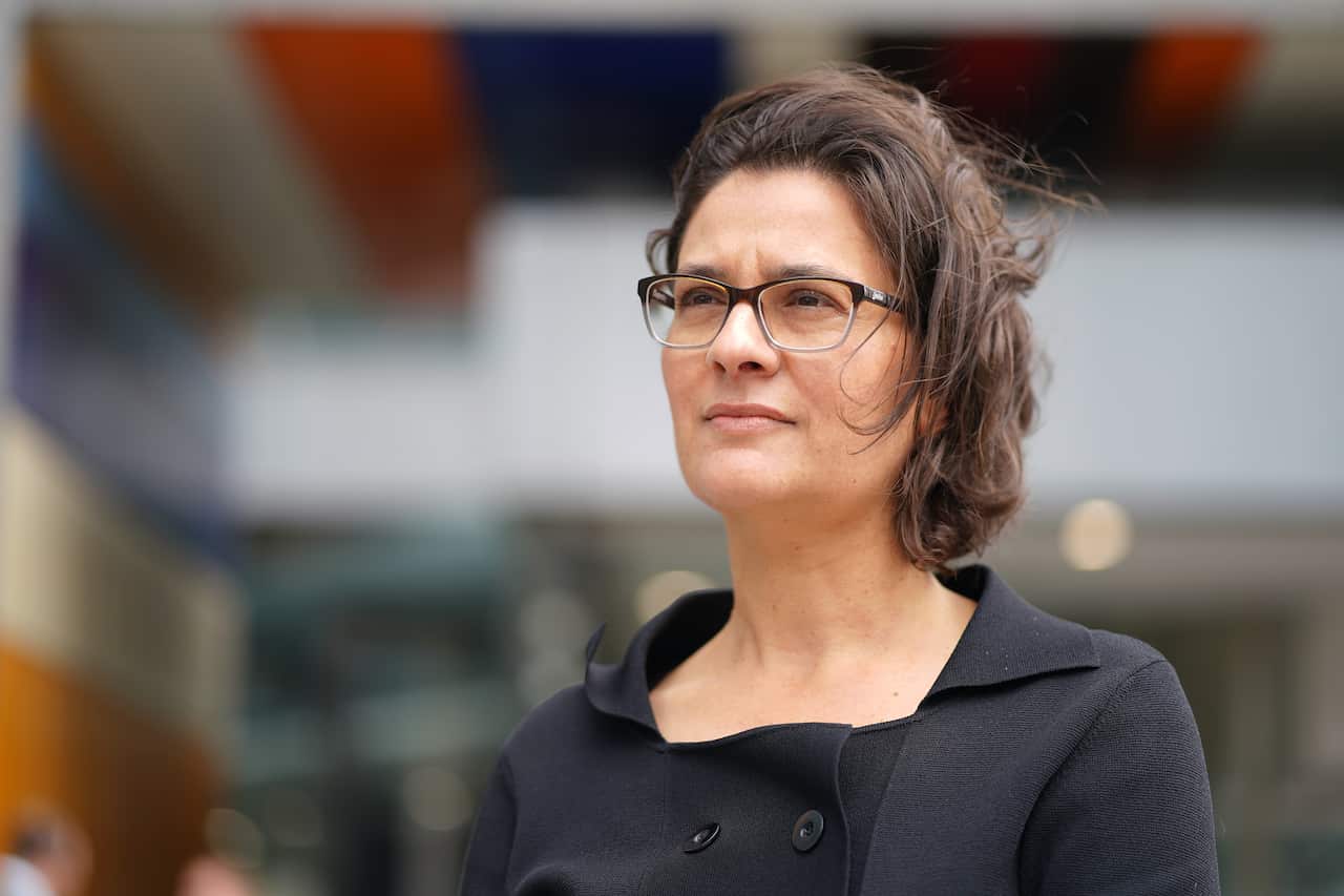 A woman with short brown hair and glasses, wearing a black coat.