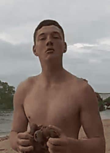 A shirtless teenage man stands in front of a river.