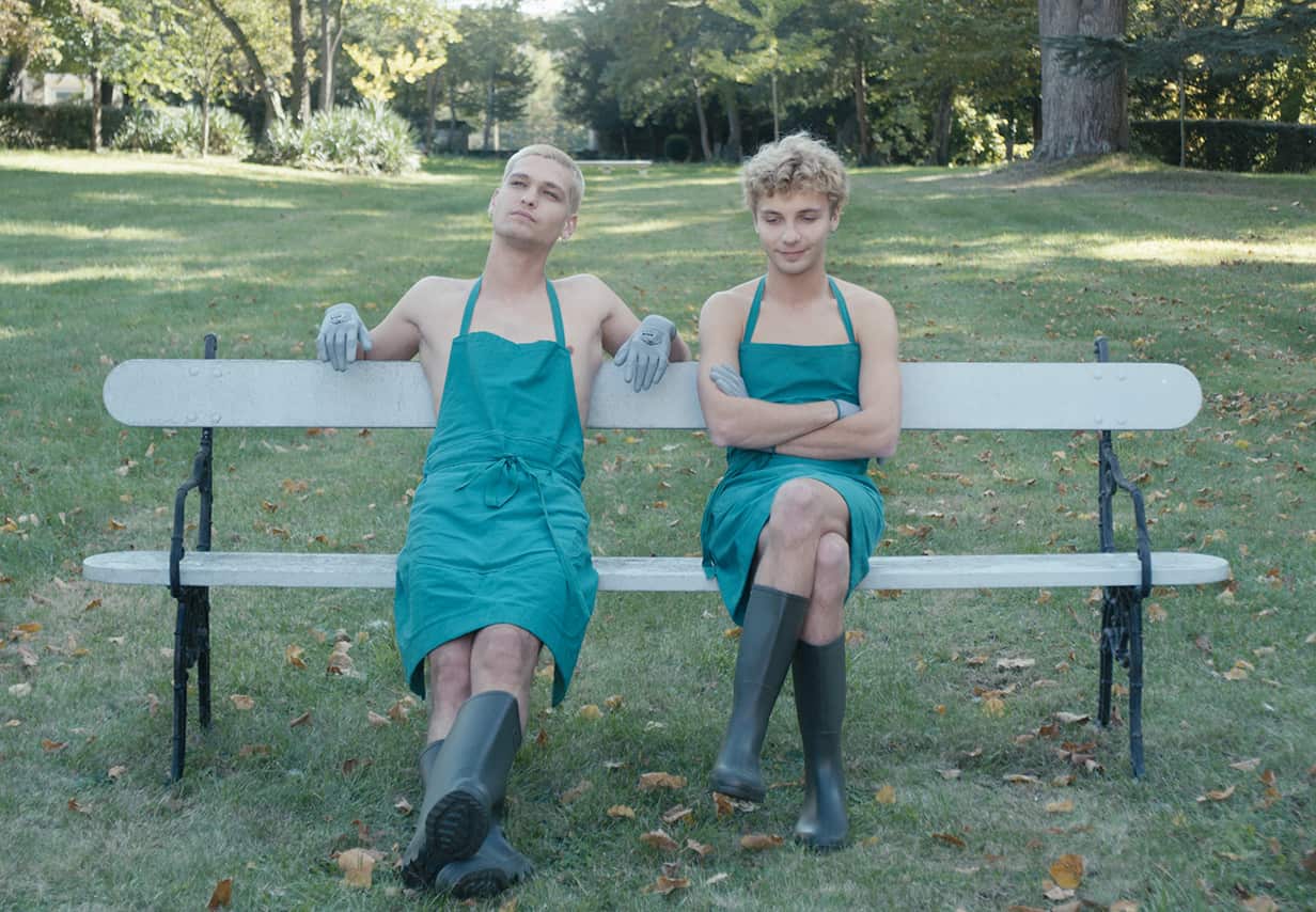 Two young people wearing aprons and gumboots (and probably nothing else) sit on a garden bench. 