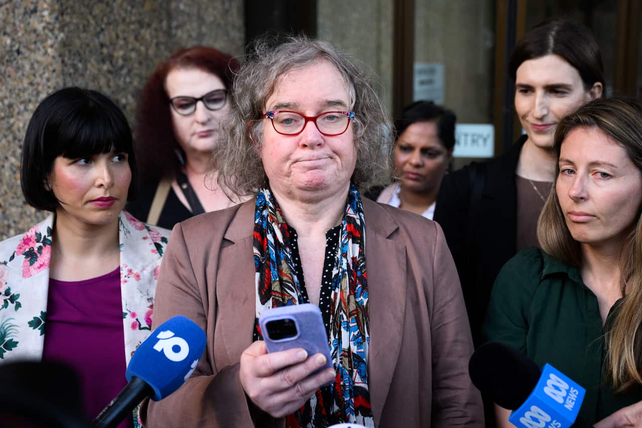 A transgender woman holding a mobile phone in front of microphones outside. There are other women around her