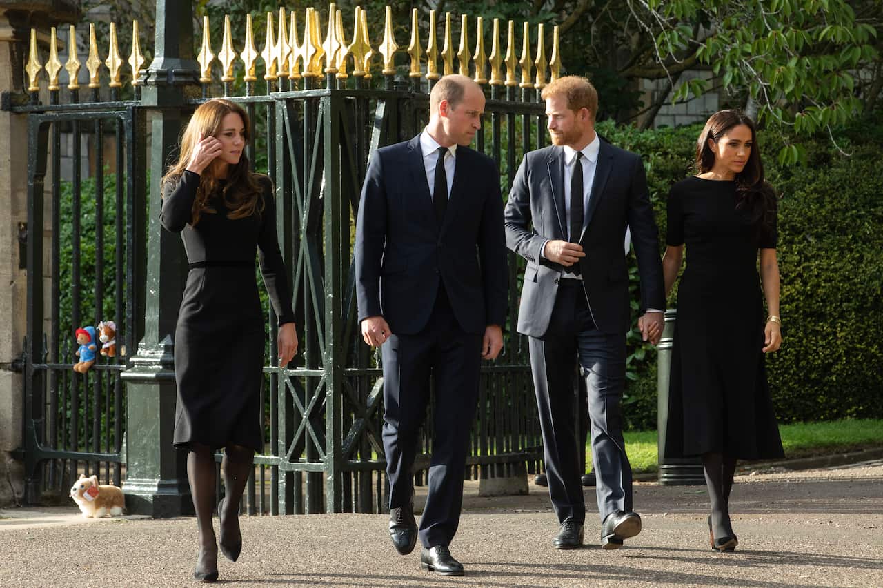 Four people walk alongside each other outside gates, two women in black dresses and two men in suits. 