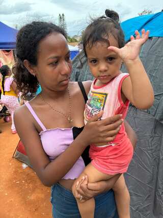 A young woman holds a young girl in her arms, who waves at the camera. They stand on dirt in front of tents.