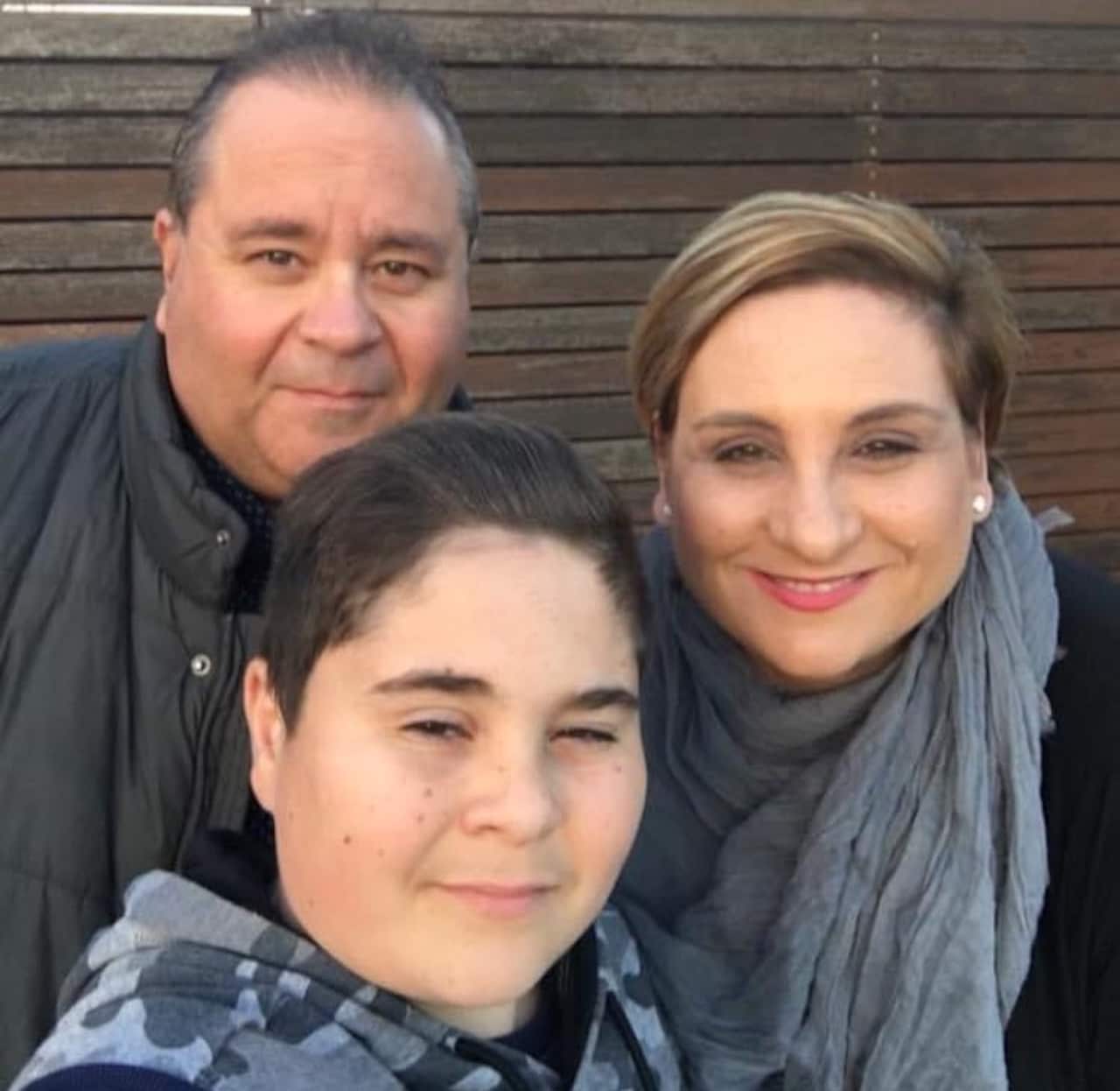 A boy wearing a grey camouflage jumper stands in front of a man and woman, his parents.