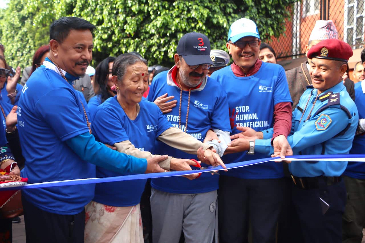 A woman in a blue shirt cutting a blue ribbon. There are other people next to her in the same shirt, and trees behind them.