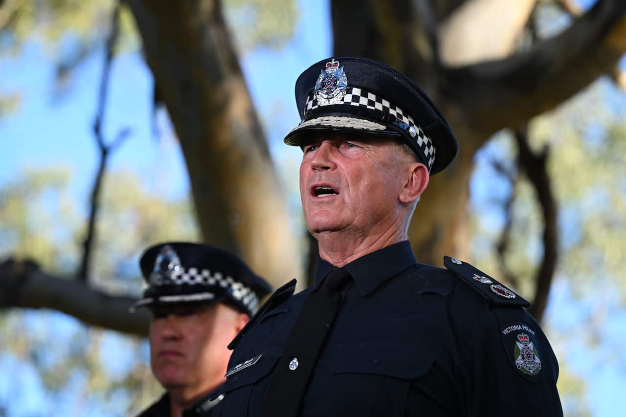 A police officer speaking in front of a tree. Another officer is next to him.