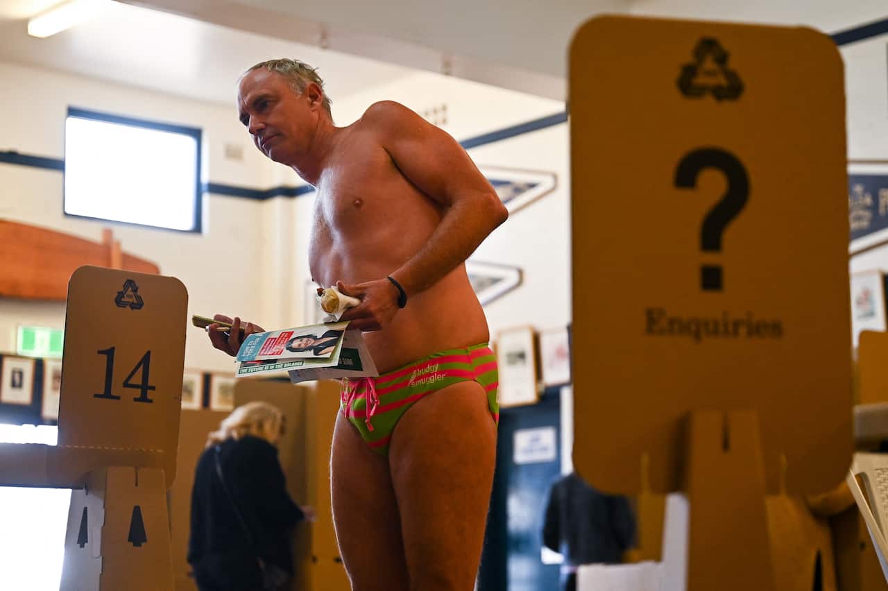 A man wearing a green and red speedo stands among several cardboard ballot boxes. 