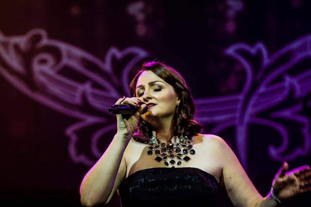 A woman in a black strapless dress holding a microphone in her right hand and singing
