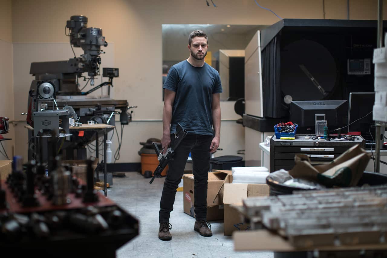 A man wearing a dark blue t-shirt and black jeans stands in a room. He is holding a black gun down by his right side.
