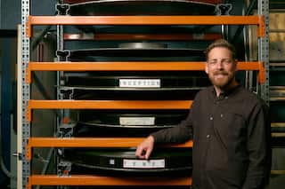 A smiling, bearded man stands next to a large orange storage rack holding massive horizontal IMAX film reels labelled "Inception" and "Tenet."