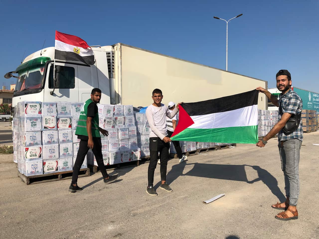 Two men holding up a Palestinian flag in front of aid trucks