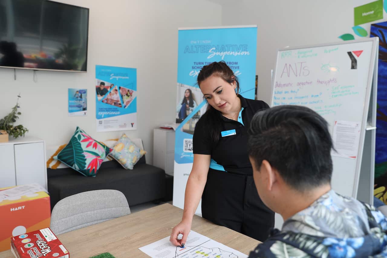 A woman standing by the desk of a student, pointing to and explaining a document on the desk.