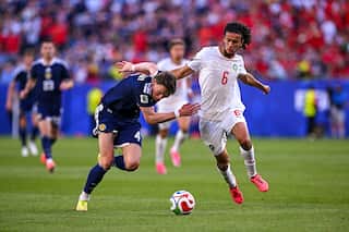 A footballer wearing a white shirt and shorts battles for possession of the ball against an opposing player.