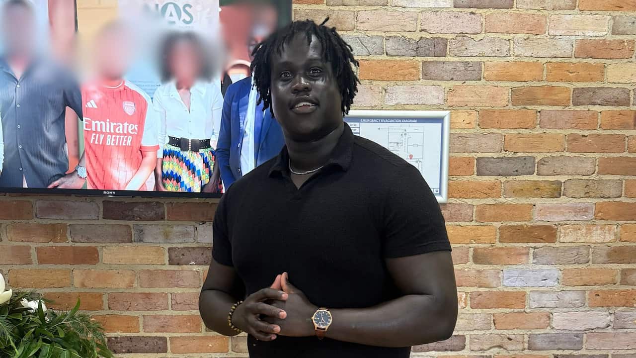 A man with dreadlocks and a black polo shirt stands in front of a brick wall, his hands clasped as he smiles towards the camera.