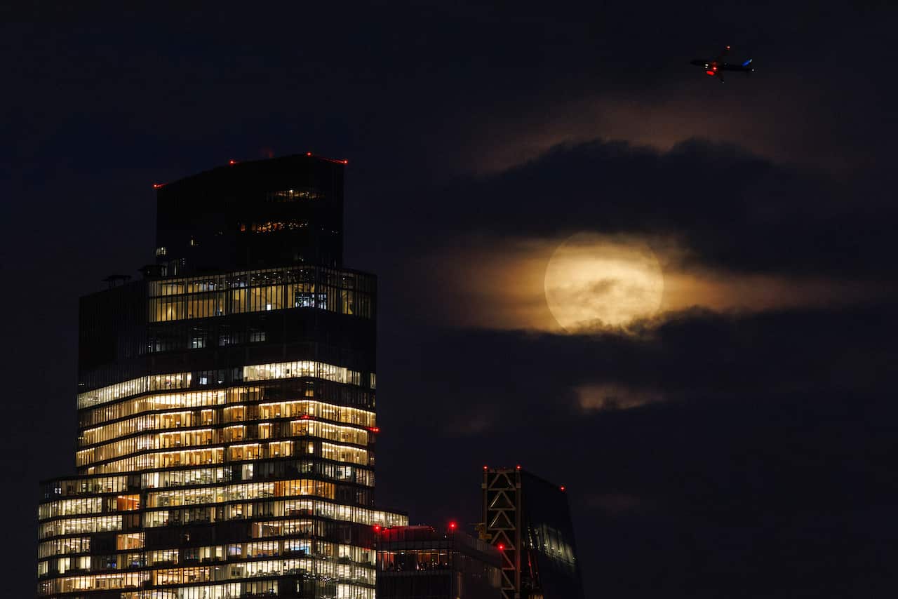 A bright, large moon behind clouds. A skyscraper can be seen next to it.