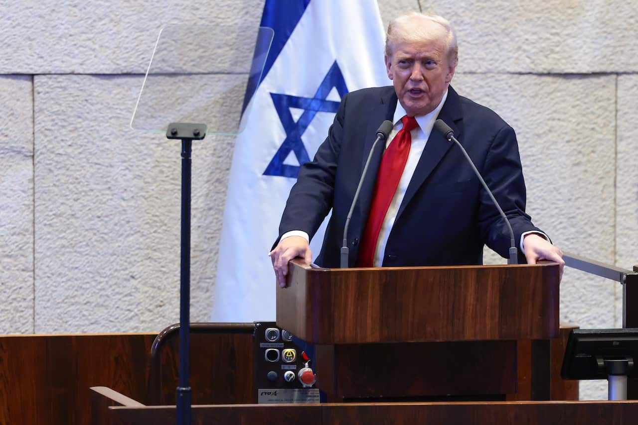 Donald Trump speaks while standing at a wooden lectern with an Israeli flag behind him.