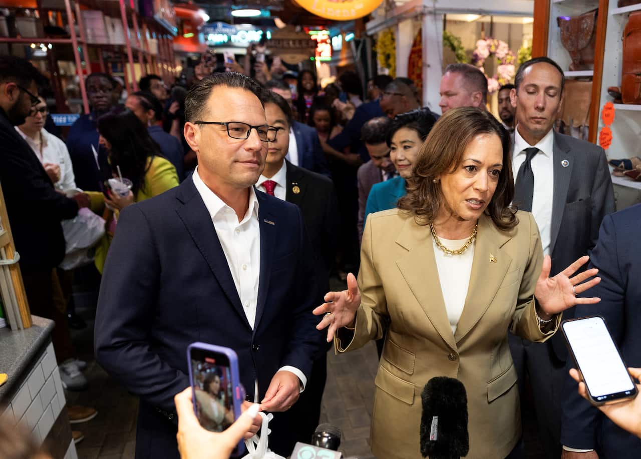A man in a navy suit and a woman in a beige suit talk to a group of reporters, huddled around them with phones.