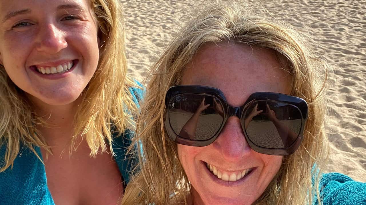a close up selfie of a young blonde woman and middle aged blonde woman on the beach with a blue towel wrapped around them