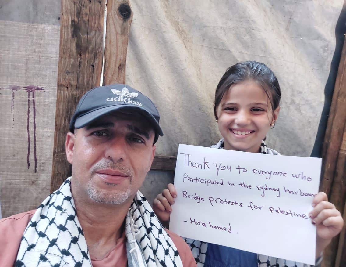 A man looks at the camera next to his smiling daughter, who holds a thank you sign.