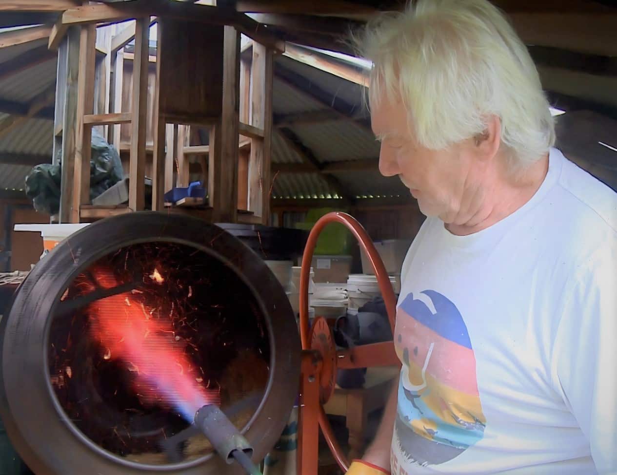 A man in a white t-shirt stands pointing a flamethrower into a barrel.