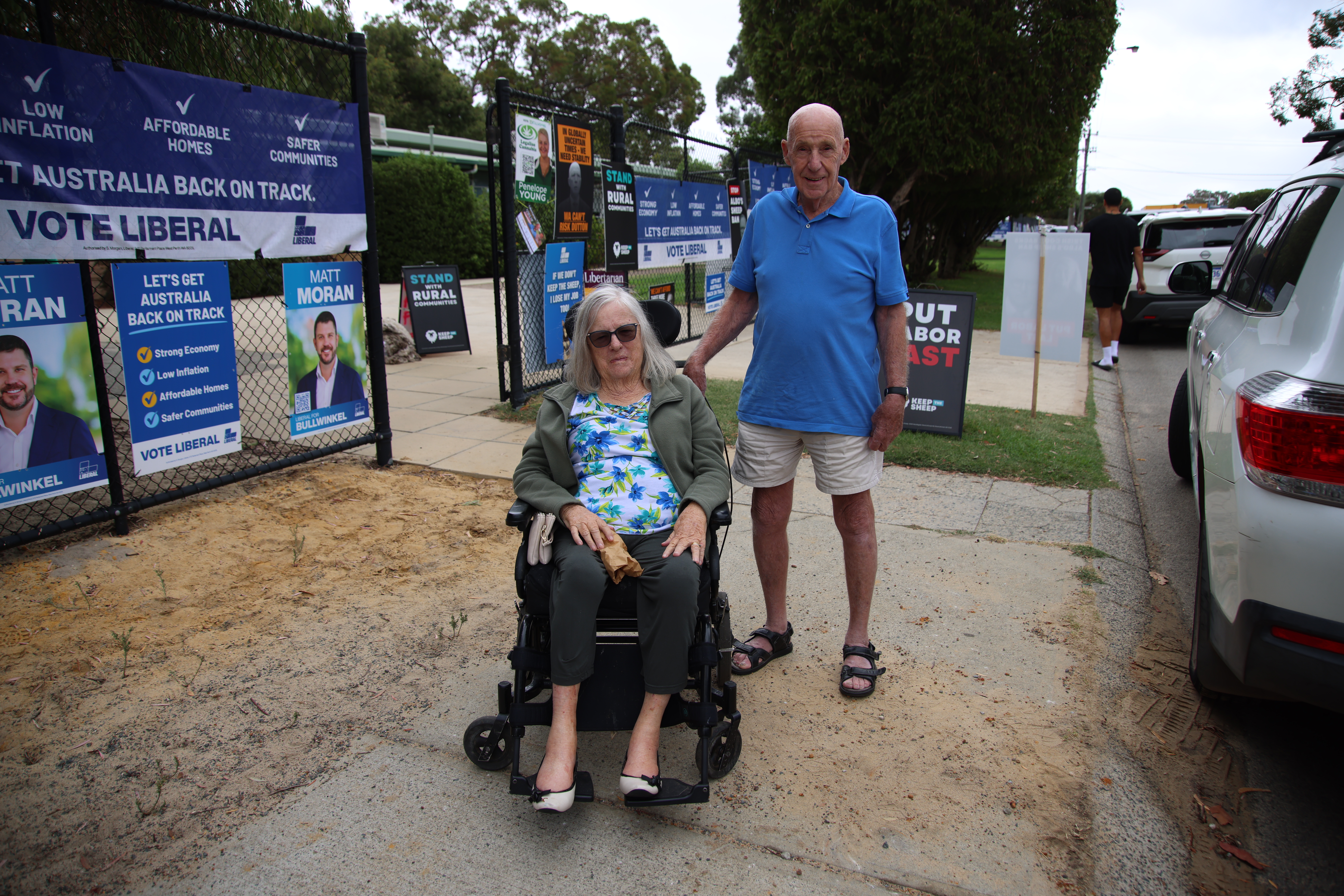 A man stands behind a woman in a wheelchair. They are both on a footpath in front of a wire fence that is covered in posted for political candidates 