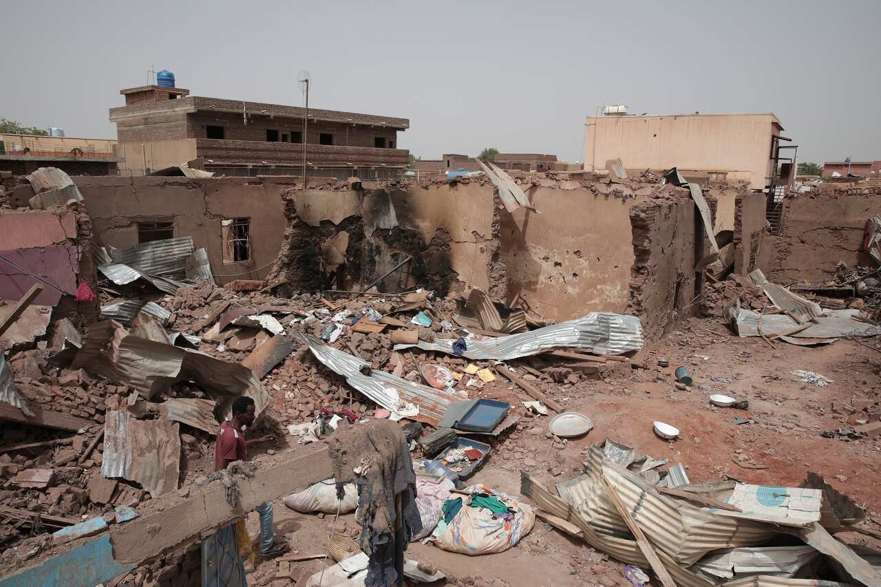 A damaged low-rise mud building and corrugated roofing on the ground.