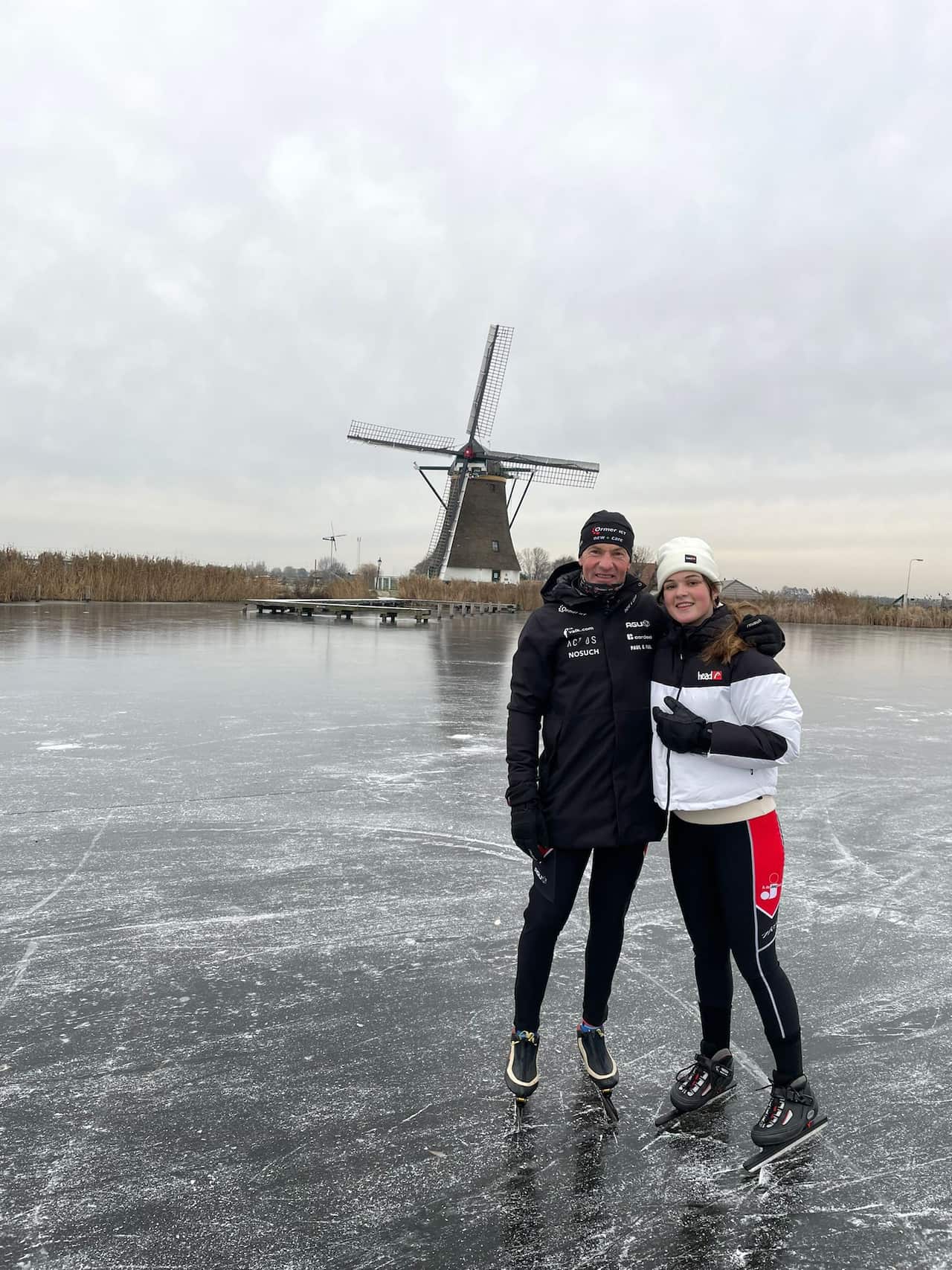 A man is hugging a young woman on a field covered by snow. In the background there is a windmill.