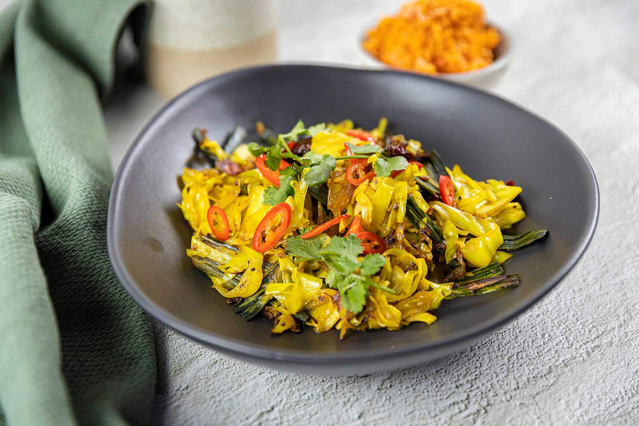 Vibrantly yellow cooked cabbage, garnished with chilli and coriander, sits in a dark bowl. 