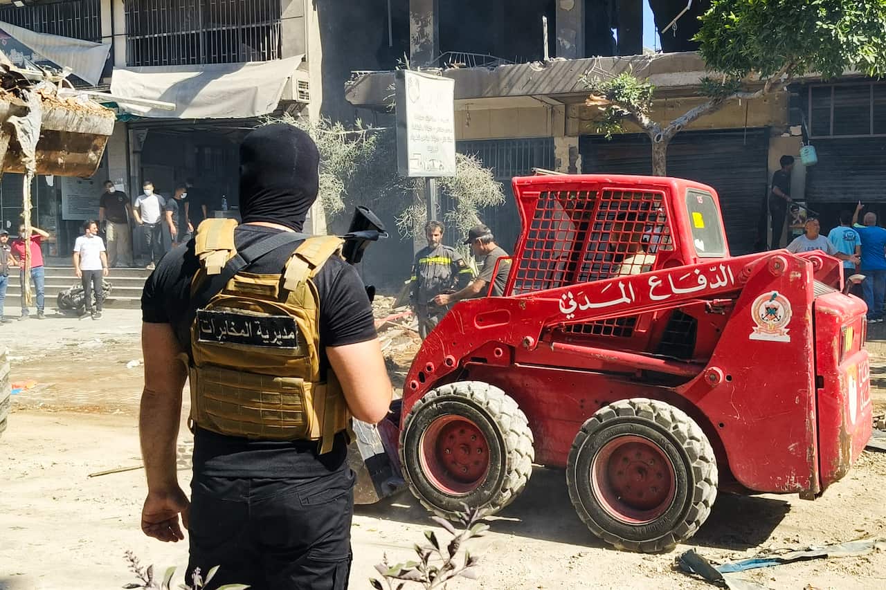 A small red bulldozer cleans up debris while an armed man wearing a black balaclava and a khaki military vest watches on.