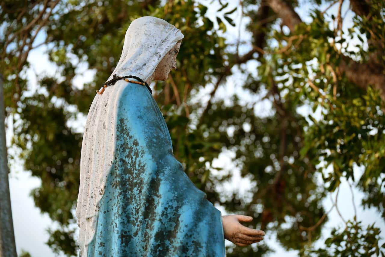 A statue of Mary outside. Someone has placed a timber rosary around her neck.