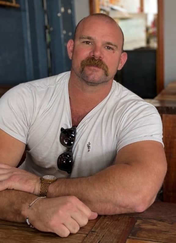 A muscular man with a prominent handlebar moustache and a shaved head sits at a wooden table wearing a white t-shirt and a gold watch.