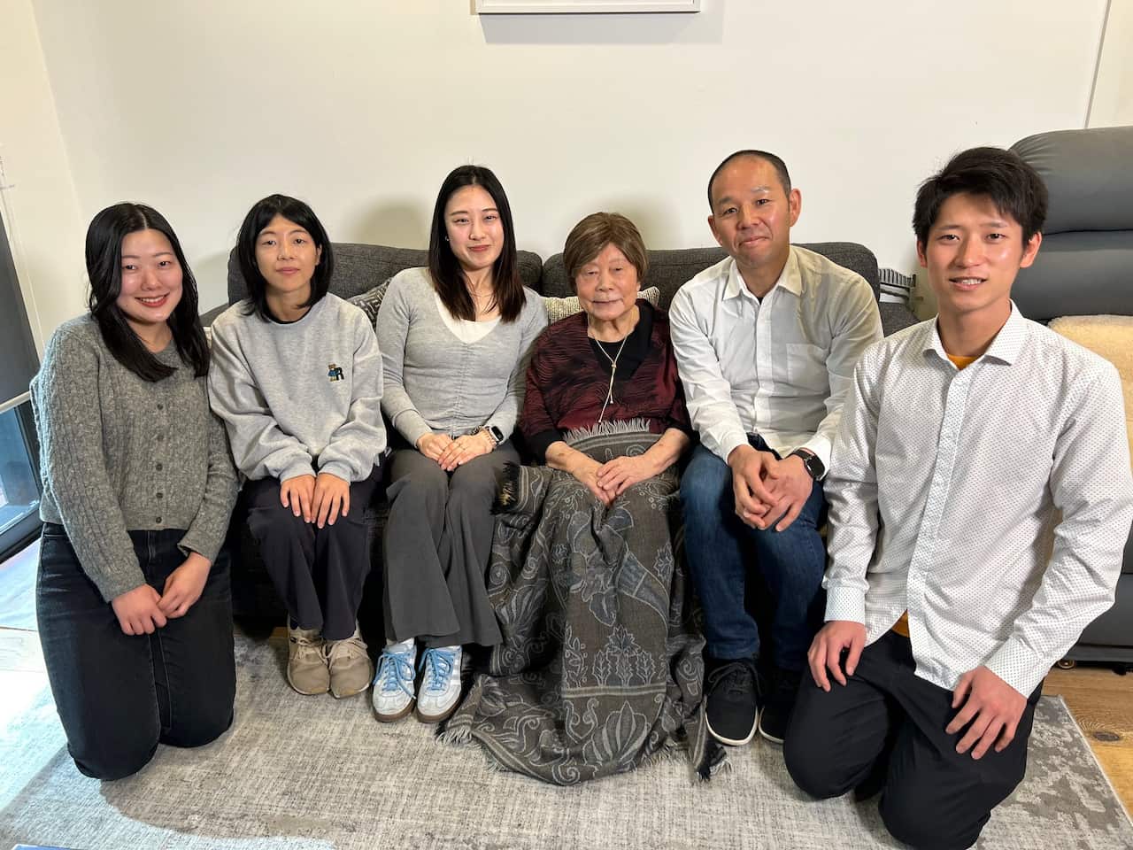 An elderly woman in a burgundy top sits on a sofa next to a group of young men and women.