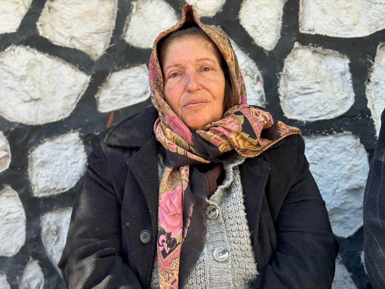 A woman sits in front of a wall, wearing a patterend scraf on her head