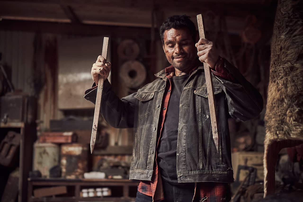 A man stands in a grubby shed holding a garden stake in each hand, looking mischievous.jpeg