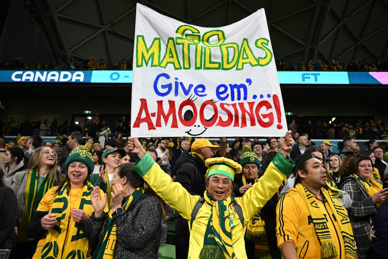 A crowd of people dressed in yellow and green. A man holds up a sign reading 'Go Matildas give em a-moosing'