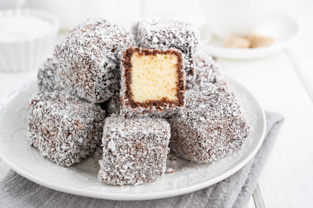 Traditional Australian Lamington cake in chocolate glaze and coconut flakes on a white plate on a white wooden background with a cup of tea. Selective focus.