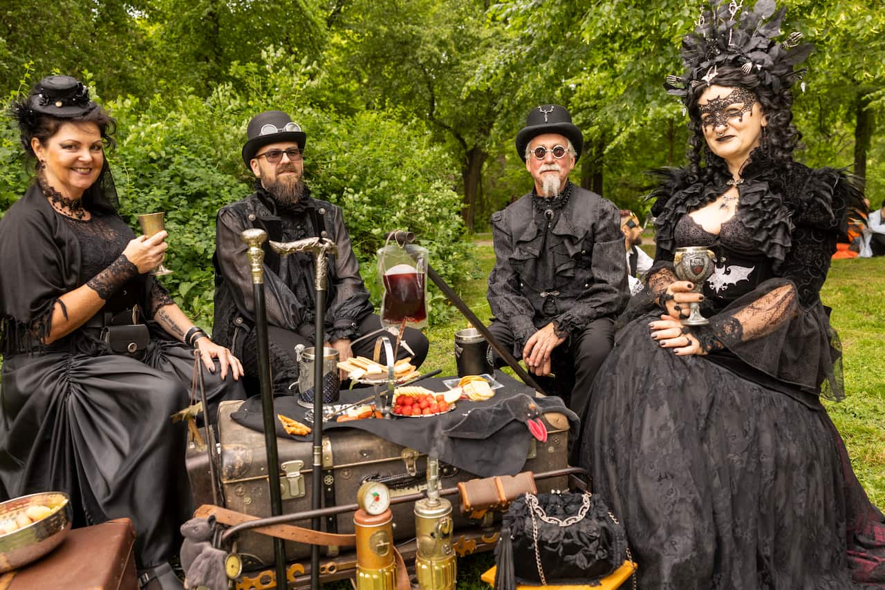 Four people dressed in elaborate, gothic and steampunk-inspired black attire enjoy an outdoor picnic complete with ornate chalices and a blood bag prop.
