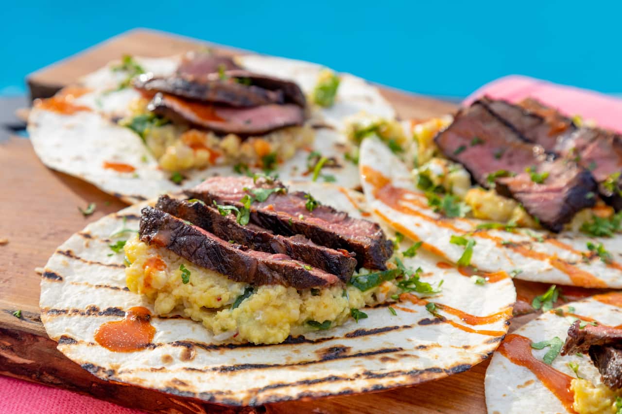 A row of flat tortillas with charry grill marks sit on a board, each with scrambled eggs, strips of steak, herbs and a drizzle of sauce. 