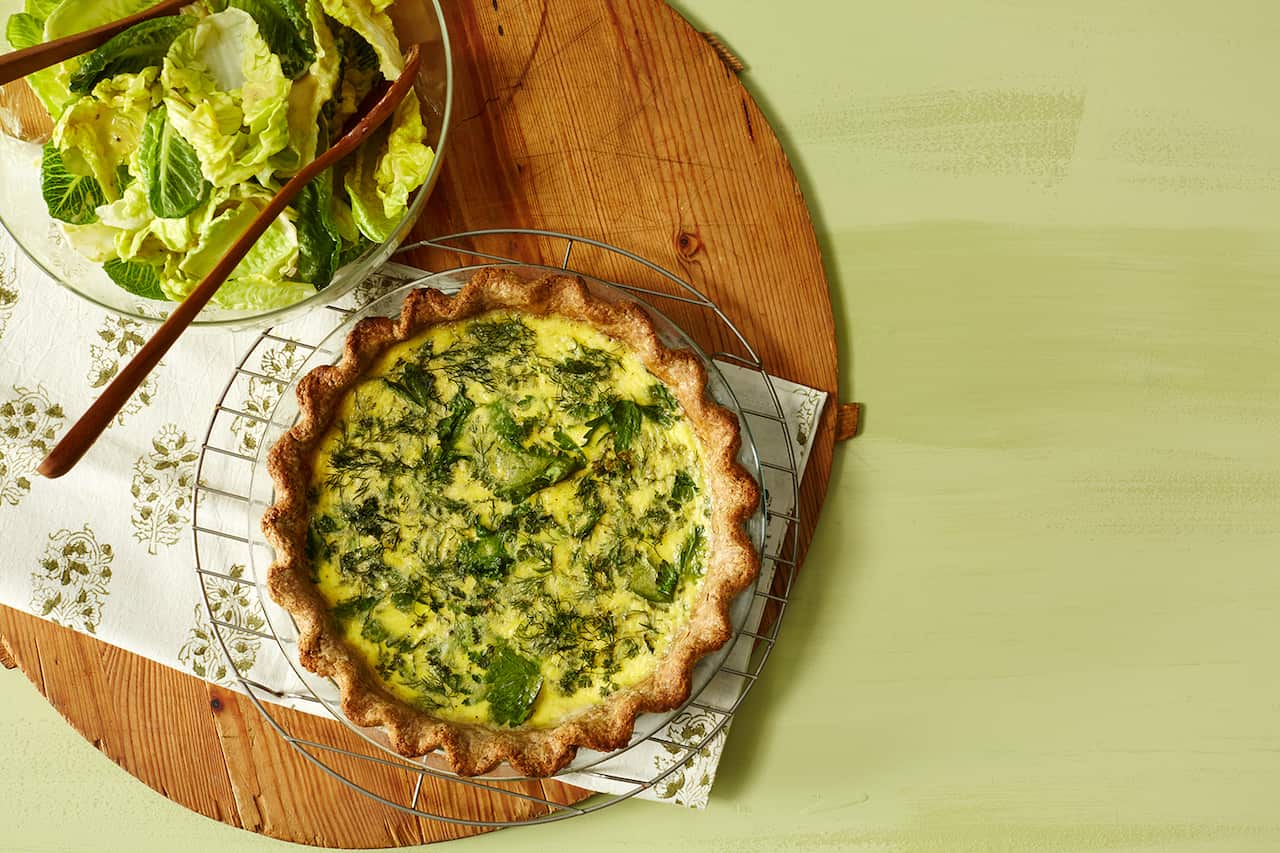 A large round woooden board sits on a green surface. On the board sits a bowl with leafy greens and beside it, a tart with fluted pastry and herby egg filling. 