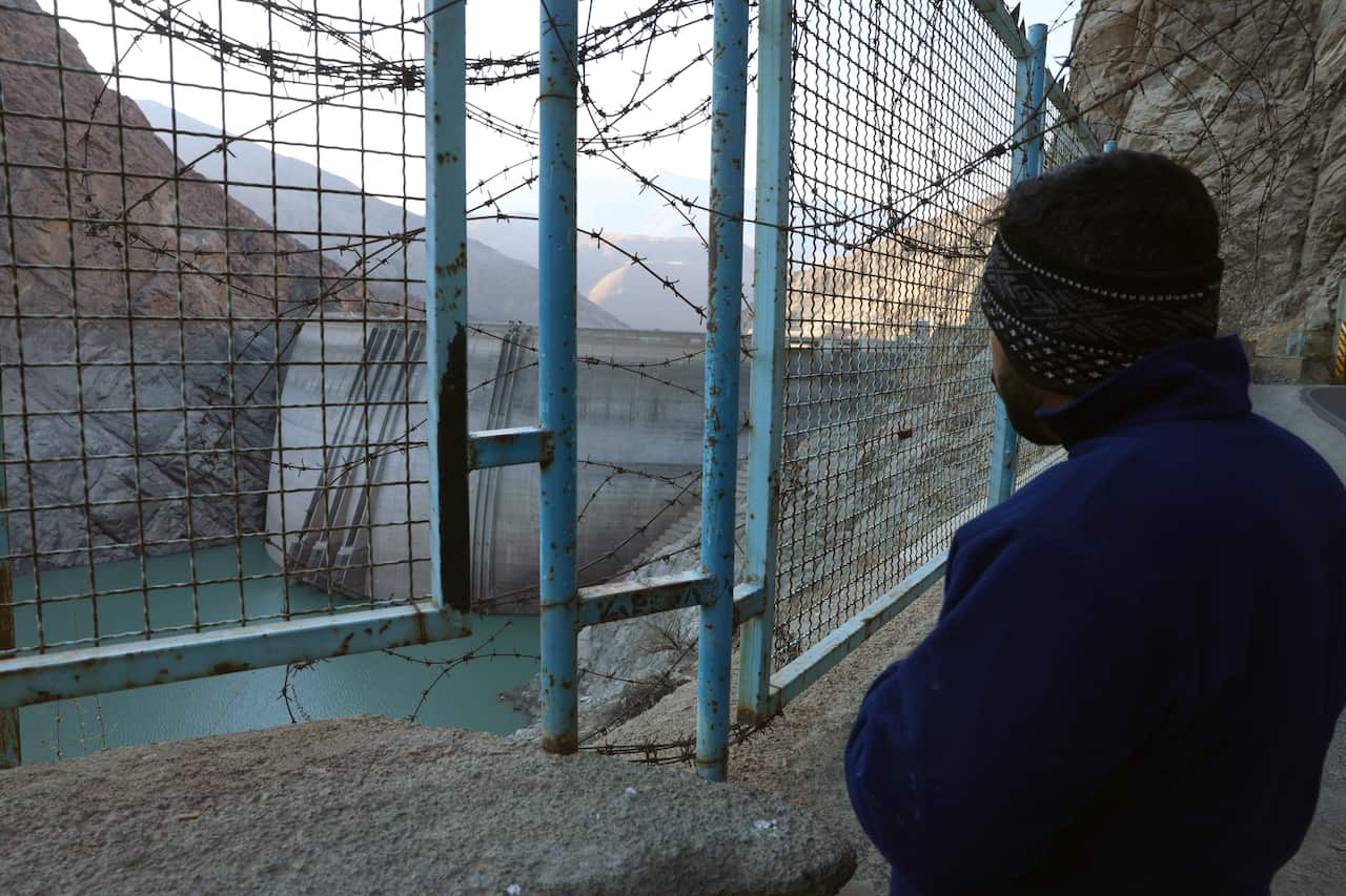 A man looking through a barbed-wire fence at a dam and a low reservoir.