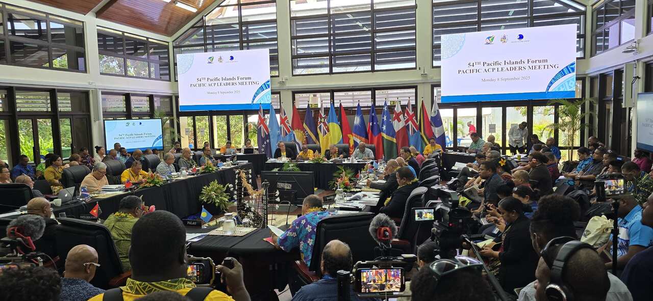 A wide-angle photo shows a large indoor meeting with a group of people seated at long tables.