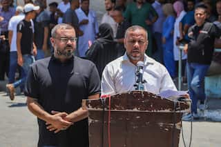 Two middle aged men stand behind a worn wooden podium. One is dressed in a dark blue shirt, the other in a white shirt. The man in the white shirt is speaking into a microphone on the podium.