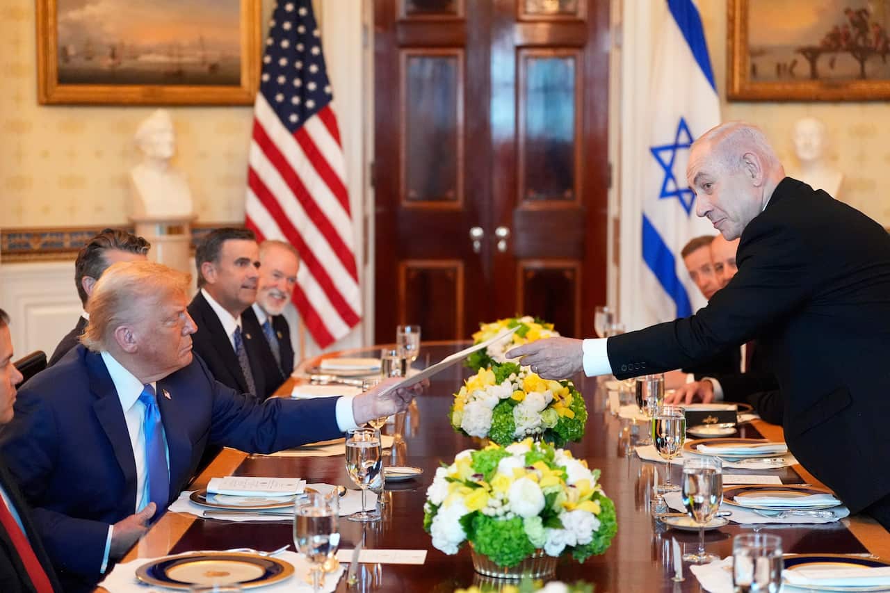 Donald Trump, sitting on the left side of a long wooden table, is being handed an envelope by Benjamin Netanyahu from across, with other delegates sitting on either side. There are also American and Israeli flags.