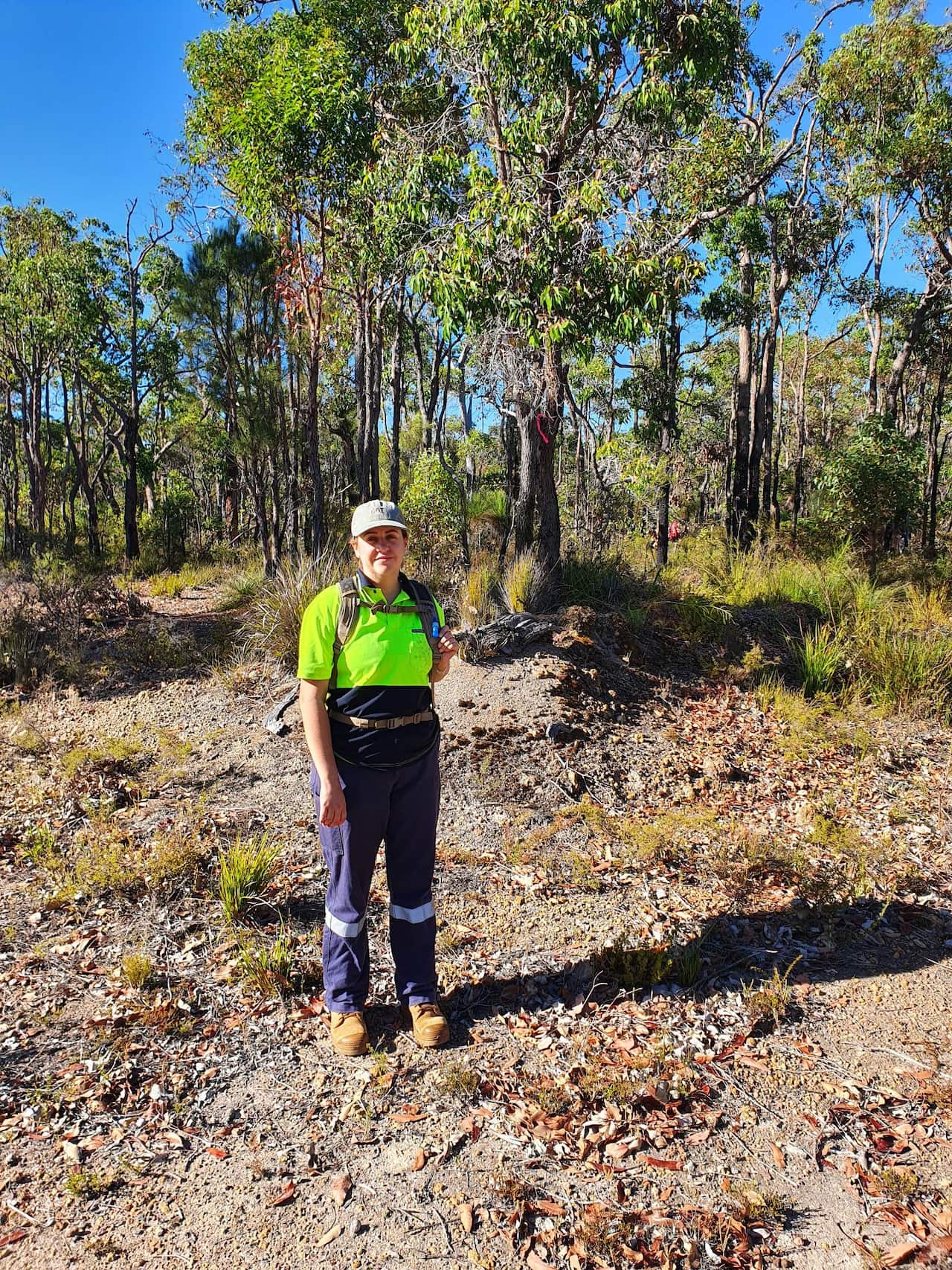 A woman in high-vis gear stands outside in the bush with a backpack on her back.