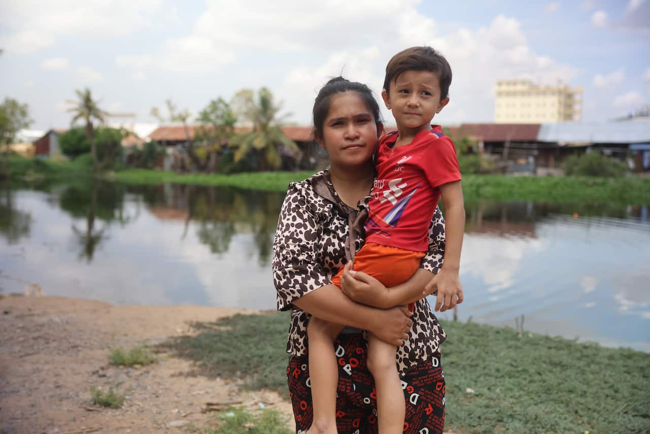 A woman holding a six-year-old boy wearing a red t-shirt and orange shorts. In the background, there's a village and a river .