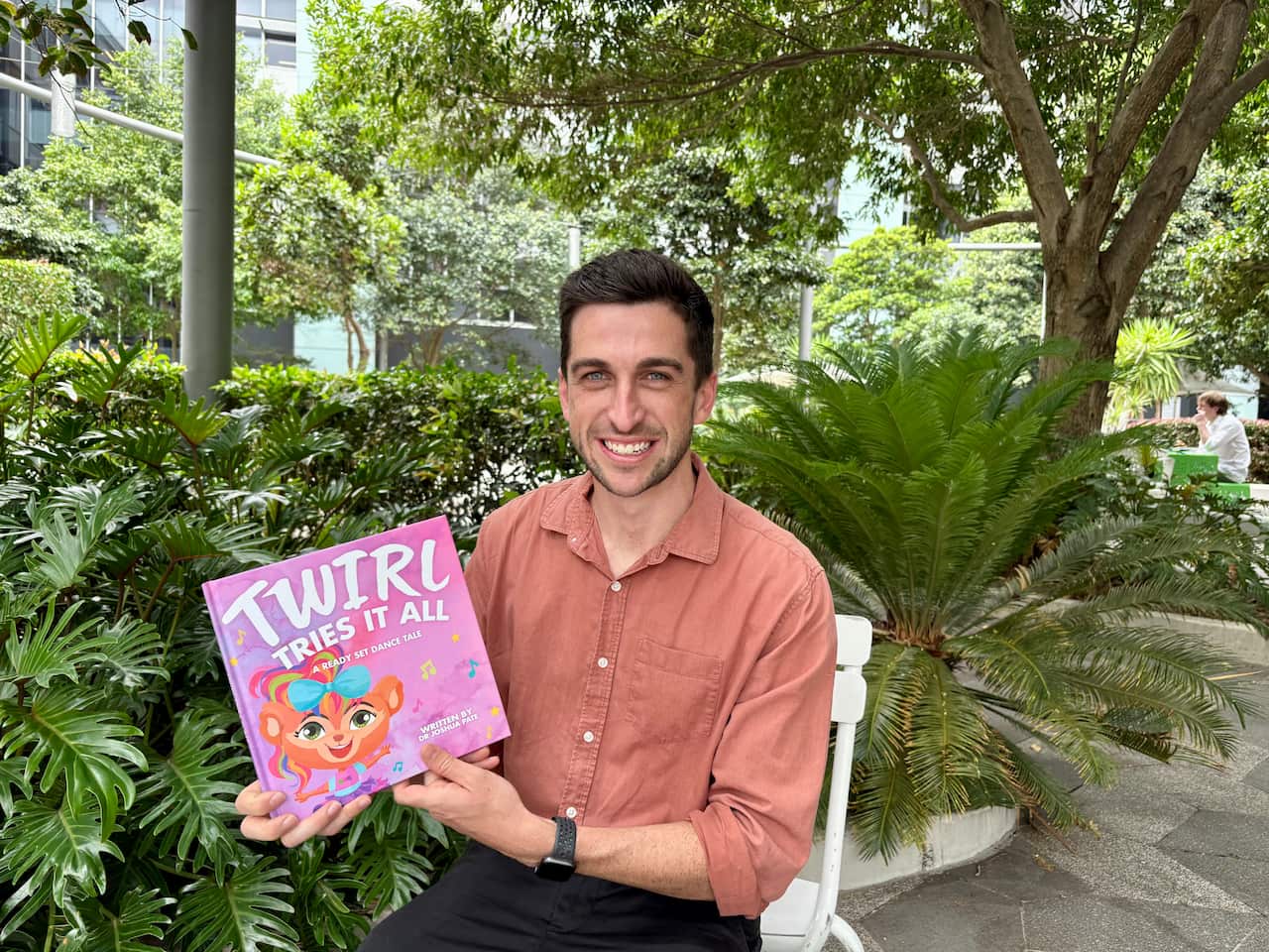 A man sitting in a plastic chair outside holds a book titled Twirl Tries It All