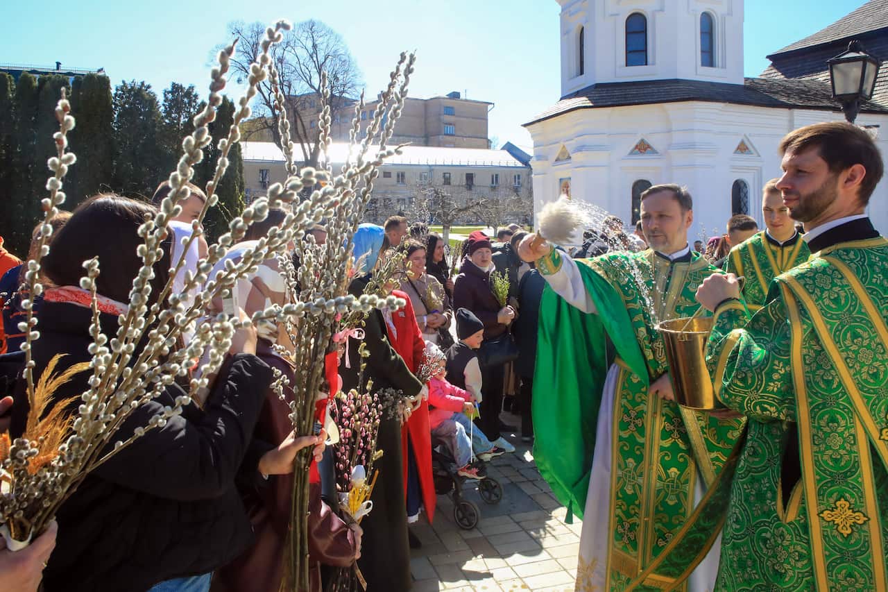 A priest blesses willow branches outside of a church as the congregation gather outside holding willow branches.